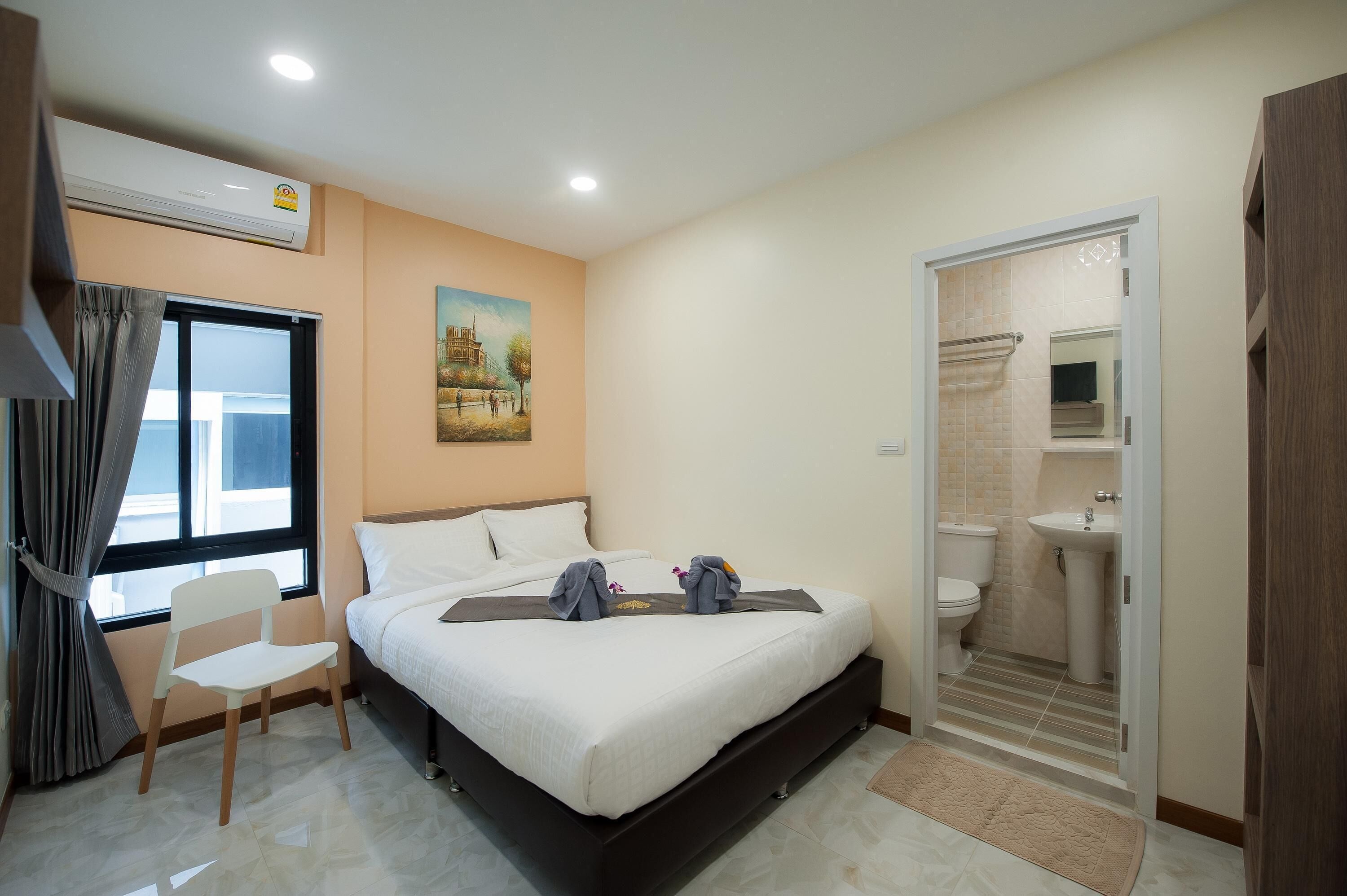 Una habitación de hotel con cama, una silla, ventana con cortinas, aire acondicionado, un cuadro en la pared y baño con lavabo e inodoro.