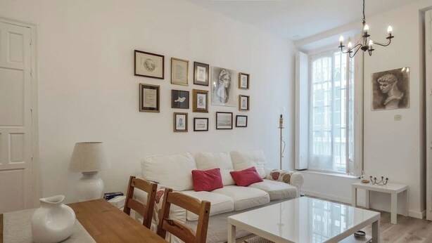 Una sala de estar moderna con un sofá blanco, una mesa de comedor de madera y una mesa de centro. Hay cuadros enmarcados en la pared, un candelabro y una ventana que deja entrar luz natural.