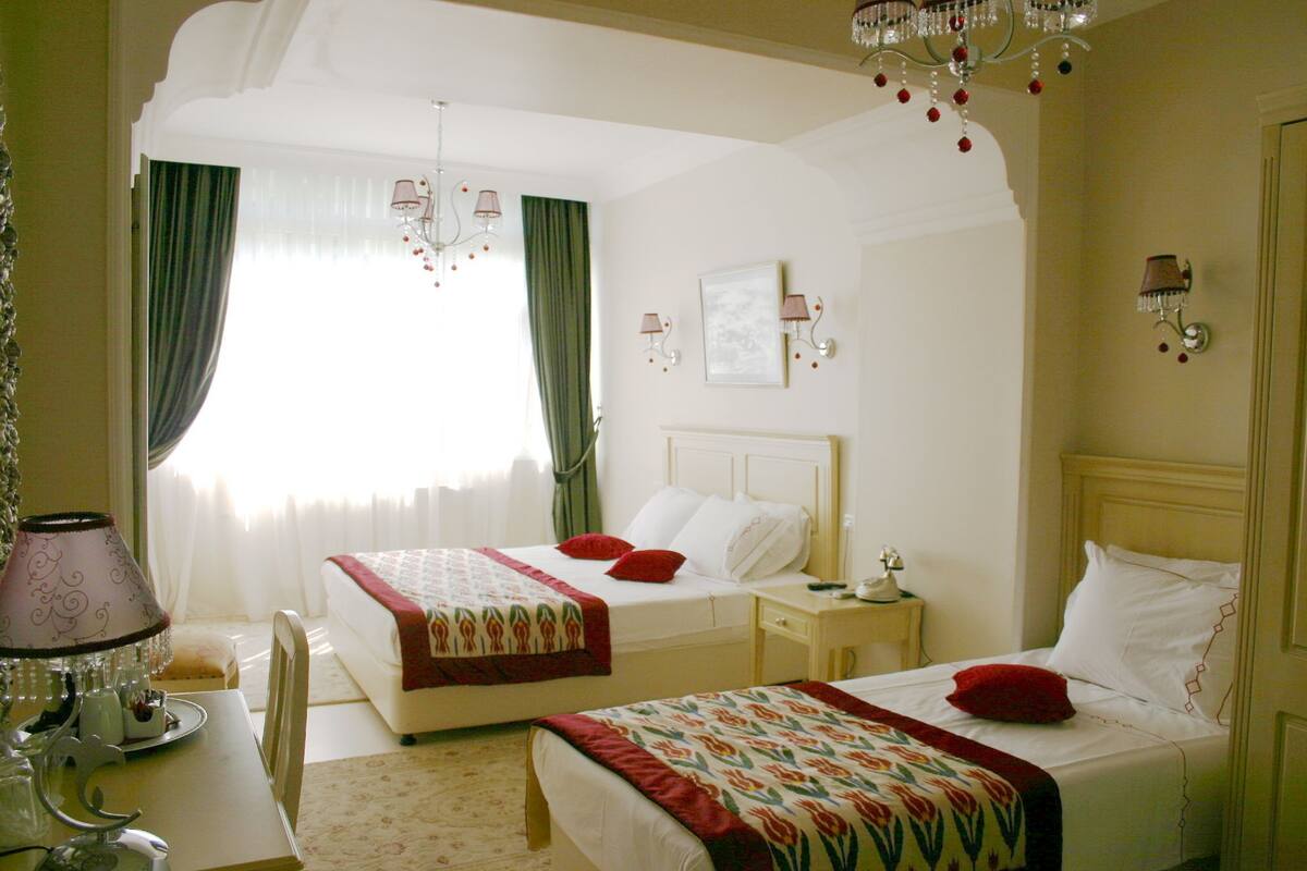 A hotel room with two beds, a chandelier, a bedside table, and a window with curtains.