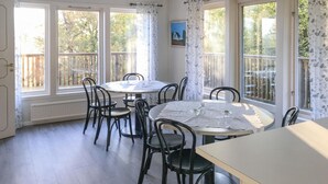 Ein Essbereich mit rundem Tisch und Stühlen, großen Fenstern mit Vorhängen sowie Blick auf Bäume und ein Haus.