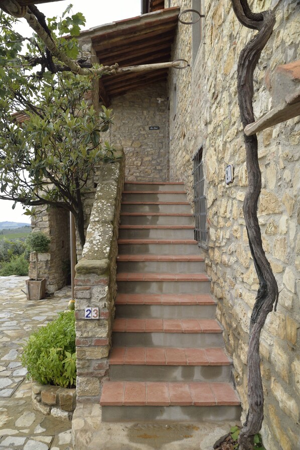 Un escalier en pierre mène à une porte en bois, et un panneau portant le numéro « 23 » est fixé au mur.
