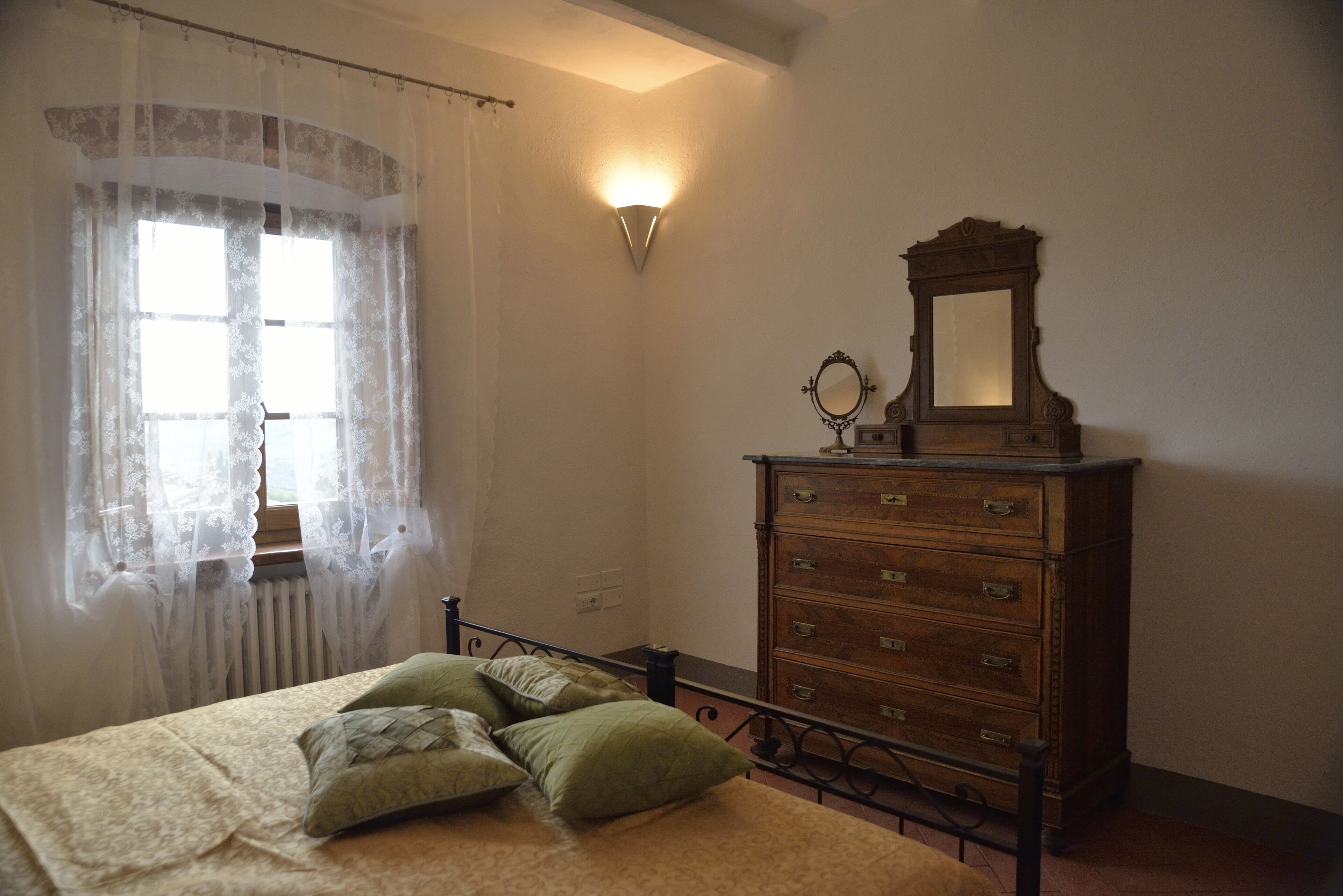 A bedroom with a bed, green pillows, a wooden dresser with a mirror, and a wall-mounted light fixture.