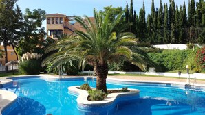 A swimming pool with a palm tree, surrounded by greenery and a building in the background.