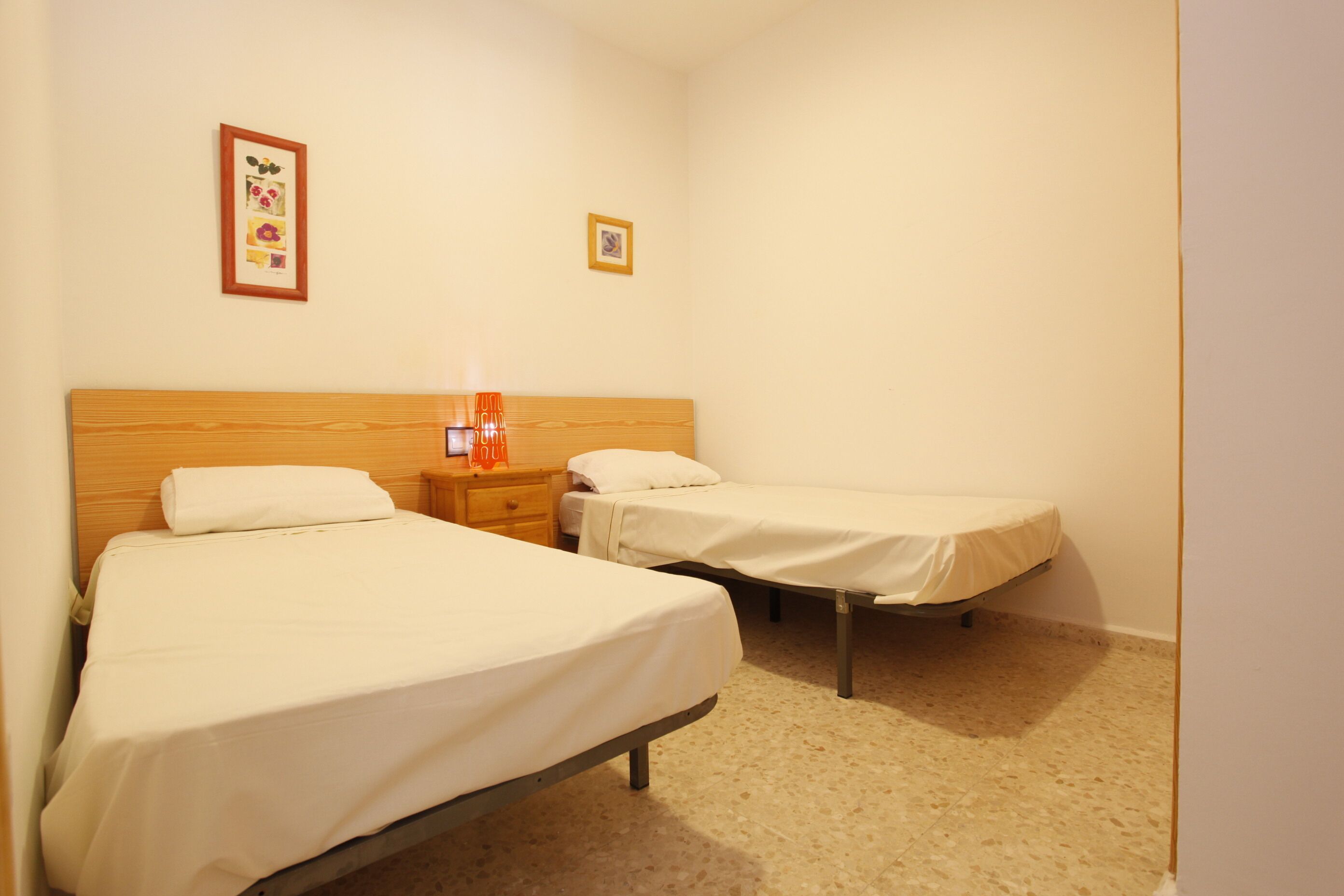 A room with two single beds, a wooden headboard, a nightstand with a lamp, and two framed pictures on the wall.