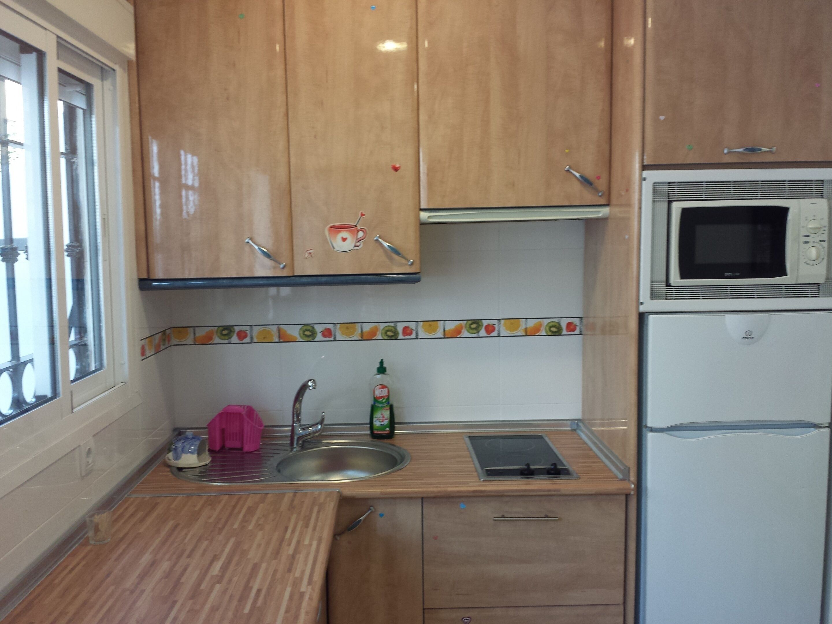 Una cocina con armarios de madera, un lavamanos de acero inoxidable, un microondas y un refrigerador pequeño.