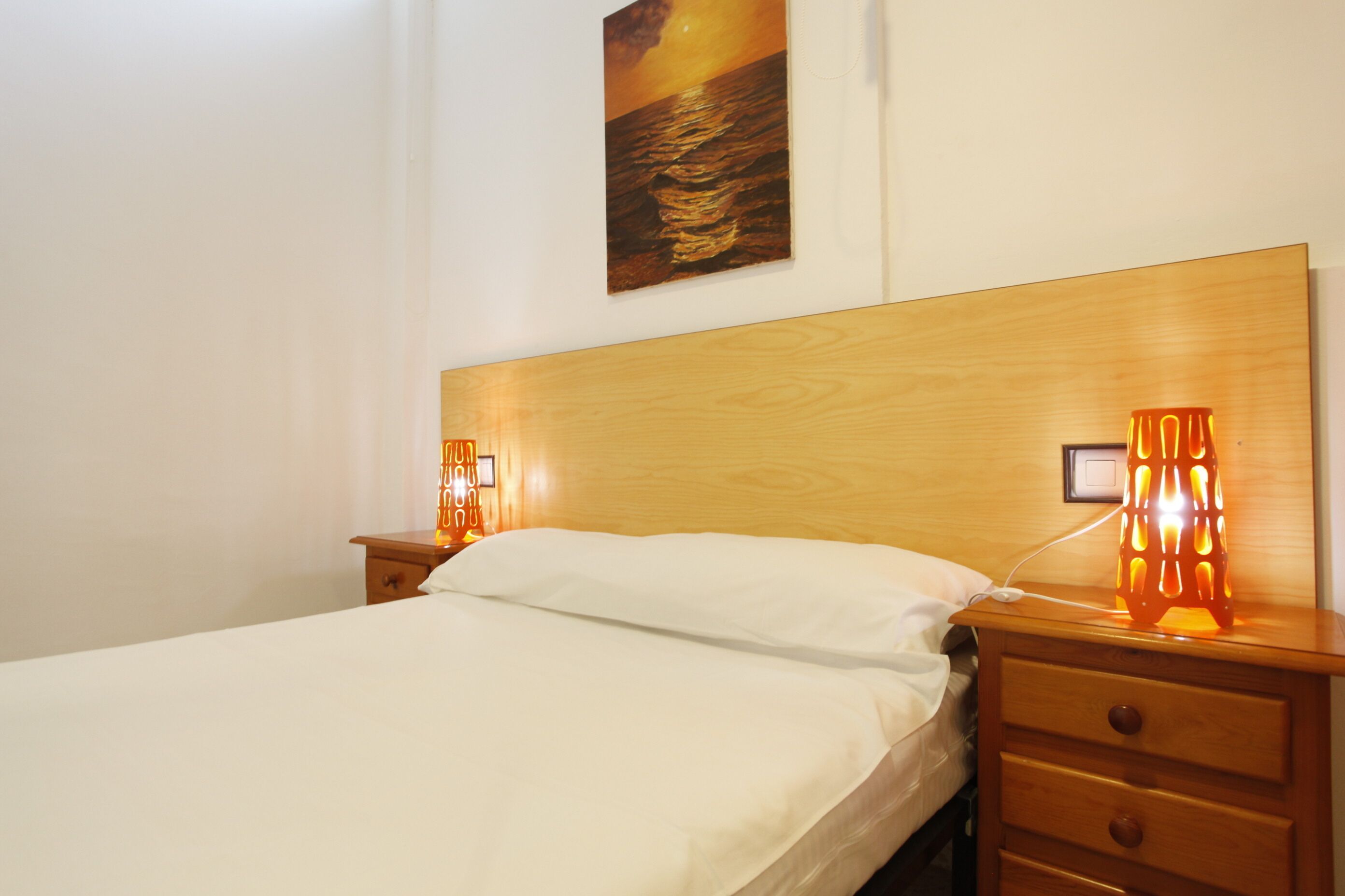 Een hotelkamer met een houten hoofdeinde, een bed met witte lakens, een nachtkastje met een lamp en een schilderij aan de muur.