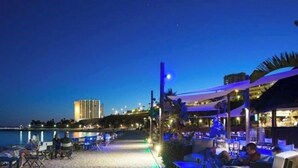 A beachfront restaurant with outdoor seating, a view of the sea, and a building in the background.