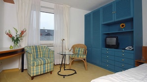 Hotelkamer met een bed, een televisie op een blauwe kast, twee stoelen, een klein tafeltje en een raam met gordijnen.