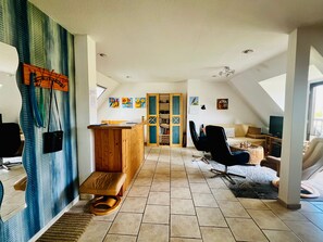 A spacious loft with a high ceiling, a wooden reception desk, a seating area with a sofa and chairs, a television, and a large window.