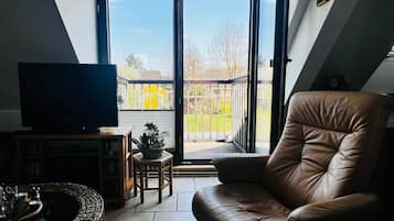 Een woonkamer met een leren bank, een flatscreen televisie, een bijzettafeltje met een plant, en uitzicht door een glazen deur op een buitenruimte.