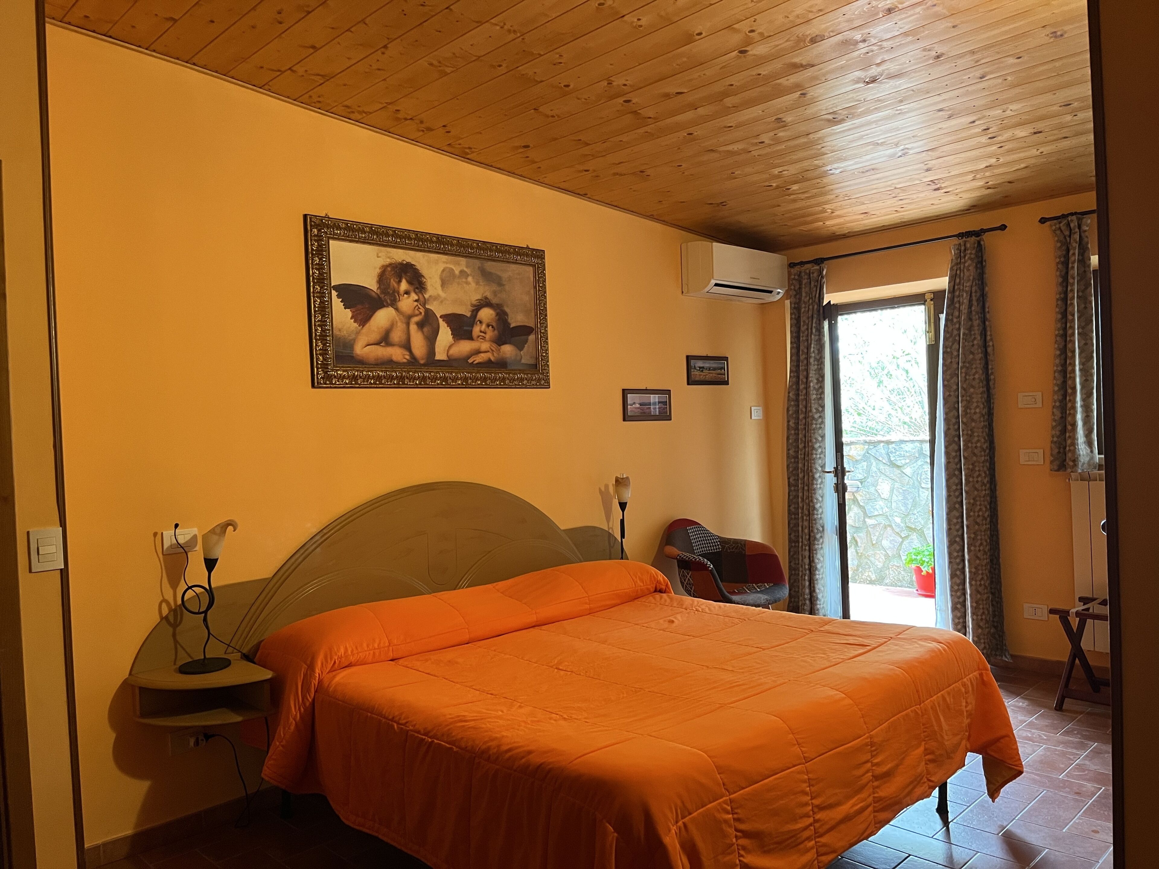 A bedroom with a bed, a painting, an air conditioner, and a view through a window.