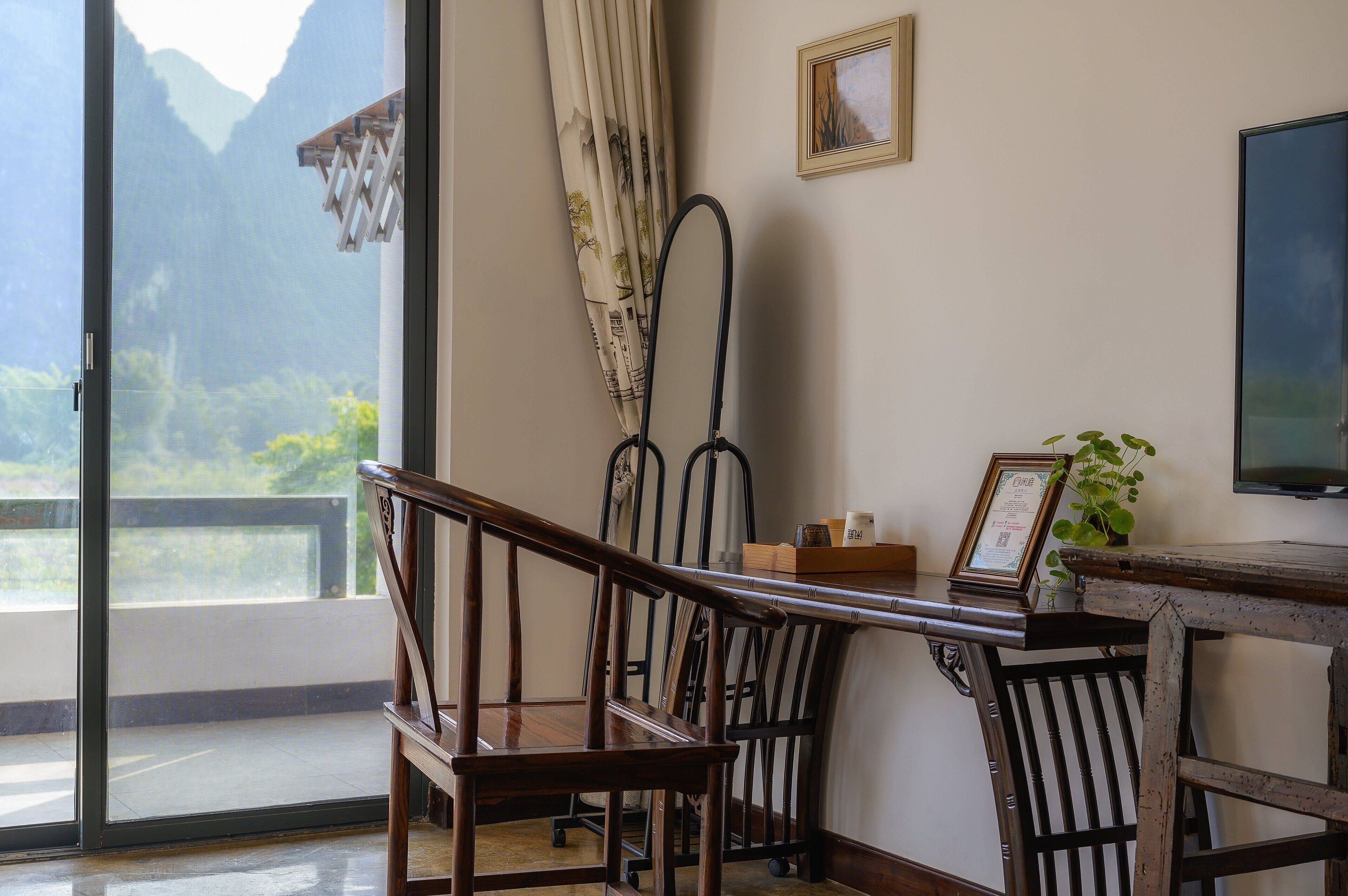 Une pièce avec une chaise en bois, une table sur laquelle est posé un cadre photo, une plante et un téléviseur fixé au mur.