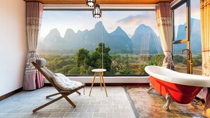 Ein Zimmer mit Holzboden, einer roten Badewanne, einem Holzstuhl, einem kleinen runden Tisch und Blick auf Berge durch große Fenster.