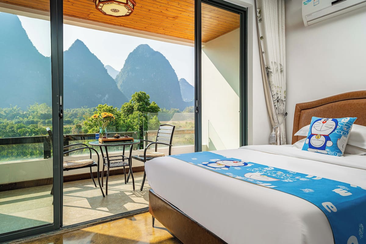 Une chambre d’hôtel avec un grand balcon, un lit recouvert d’une courtepointe ornée d’un personnage de dessin animé bleu, et une vue sur les montagnes.