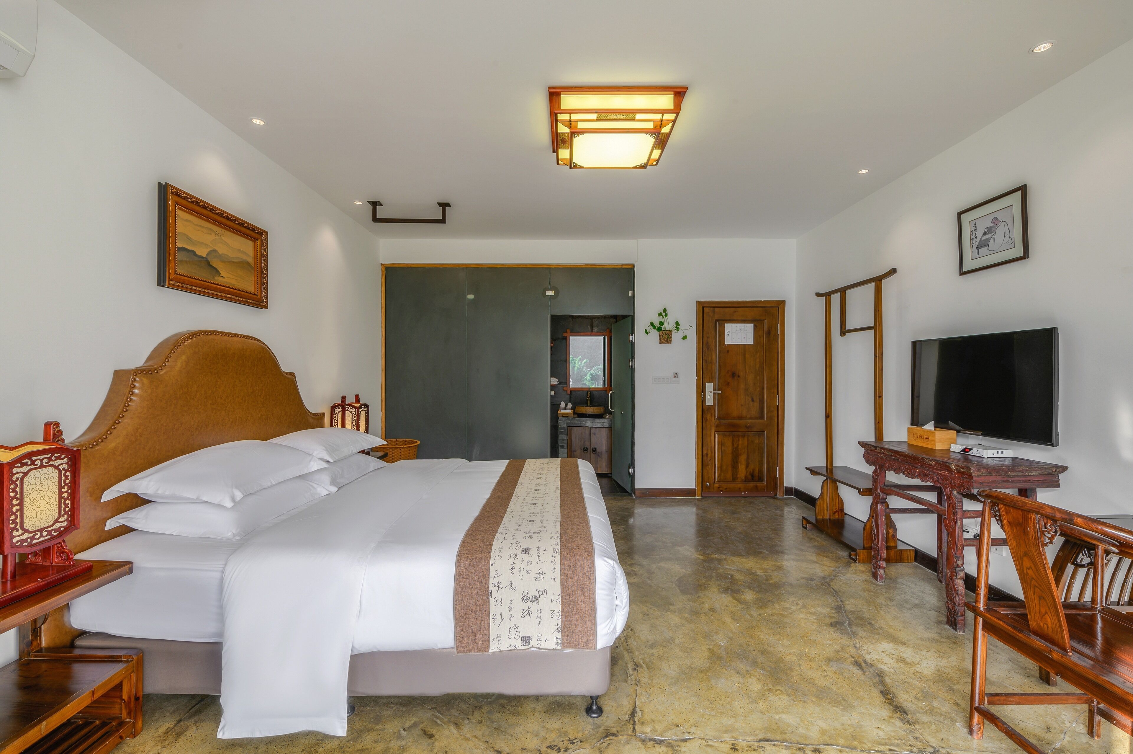 Quarto de hotel com uma cama grande, uma escrivaninha de madeira, uma televisão e um banheiro visível através de uma porta aberta.