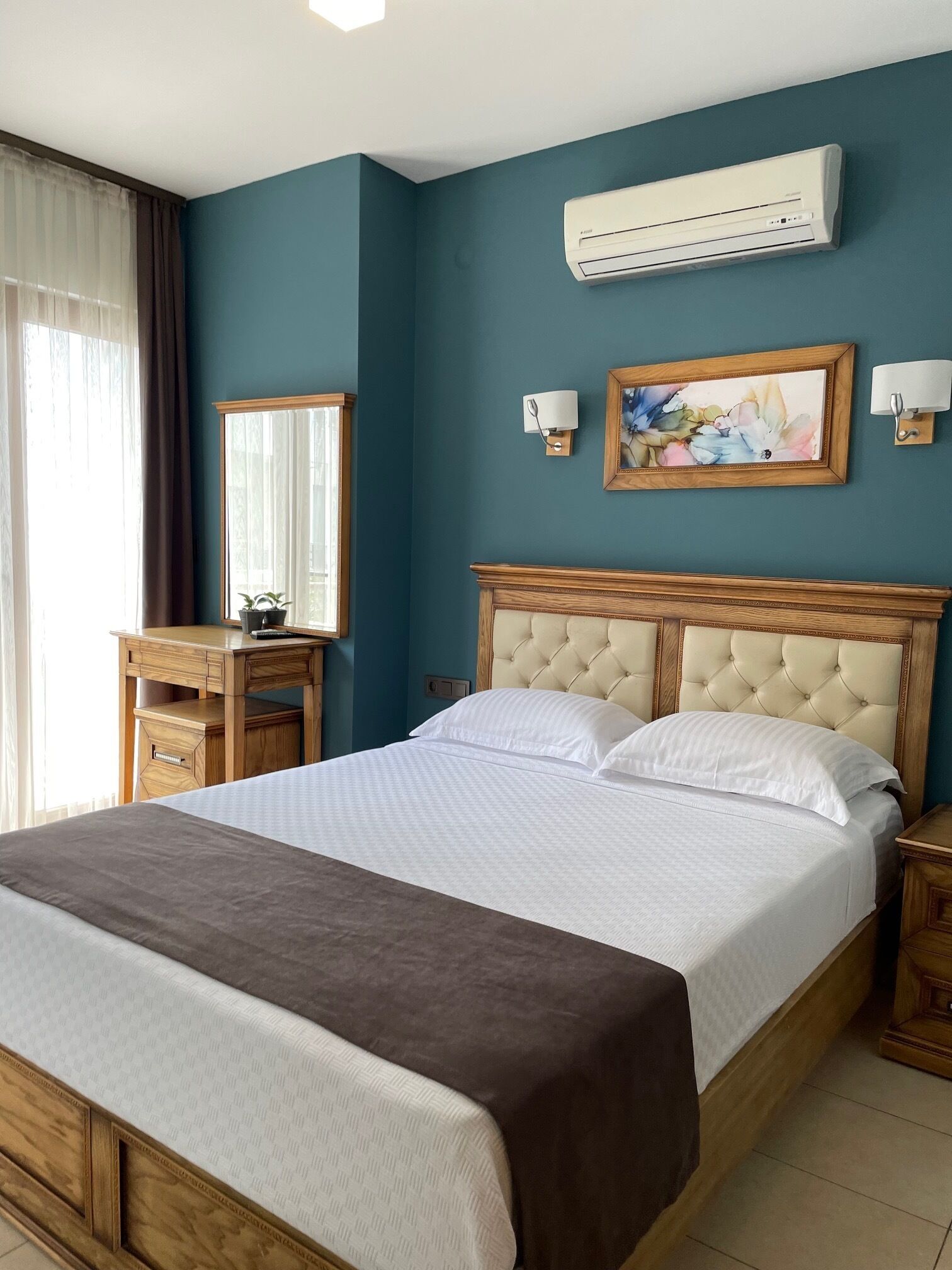 A bedroom with a bed, a nightstand, a dresser, a mirror, an air conditioning unit, and a painting on the wall.