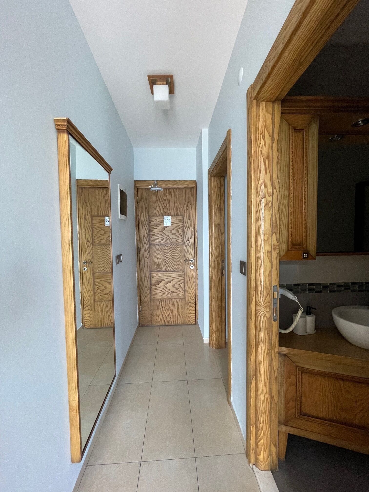 Un pasillo con puertas de madera, suelo de baldosas y un baño con lavabo y espejo.