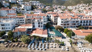 A coastal town with a beach area featuring rows of sun loungers and umbrellas, surrounded by multi-story buildings with red-tiled roofs. - Veziroglu Apart (Datça)