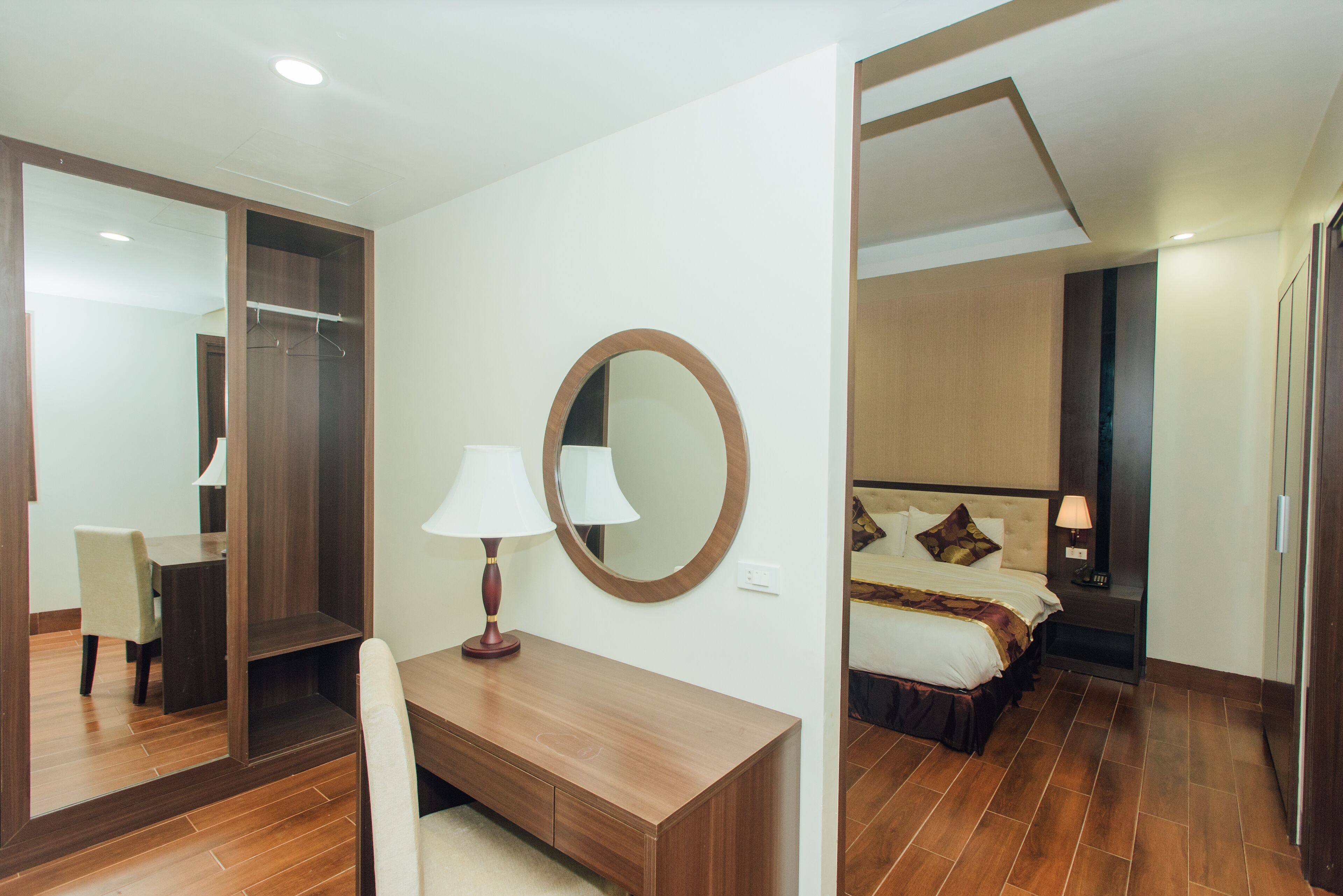Une chambre d’hôtel avec un bureau en bois, un miroir, un lit avec une tête de lit et une table de chevet avec une lampe.