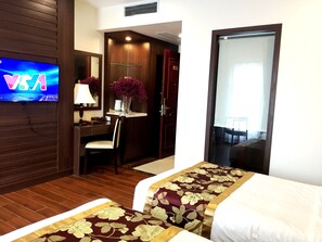 A hotel room with a TV displaying 'VIA', a desk with a lamp, a chair, a desk lamp, a vase with purple flowers, and a bed with a floral bedspread. - Hung Long hotel (Dong Nguyen)