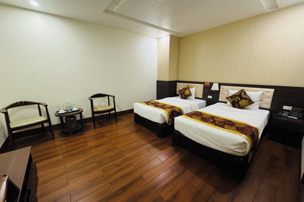 A hotel room with two beds, a wooden floor, a small table with a chair, and a nightstand with a phone.