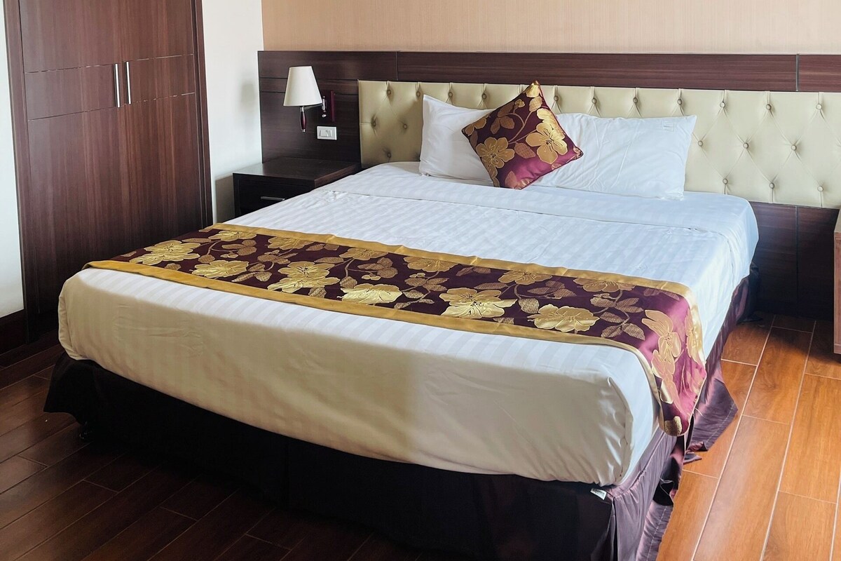 A hotel room with a large bed, a wooden wardrobe, a nightstand with a lamp, and a beige headboard.