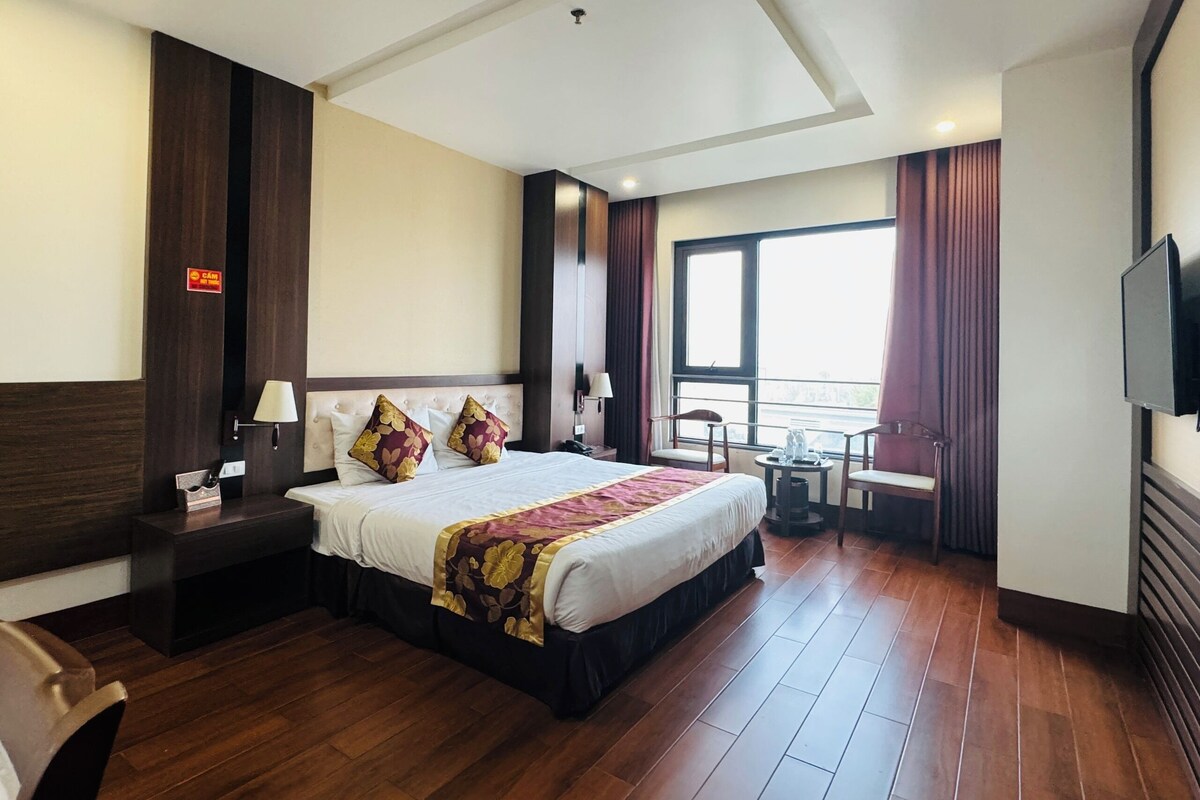 A hotel room with a large bed, bedside tables, a desk, a chair, a TV, and a window with curtains.