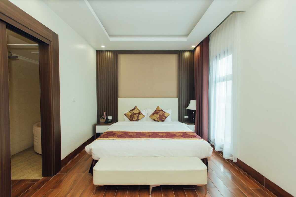 A hotel room with a large bed, a bench, a nightstand, and a bathroom visible through an open door.