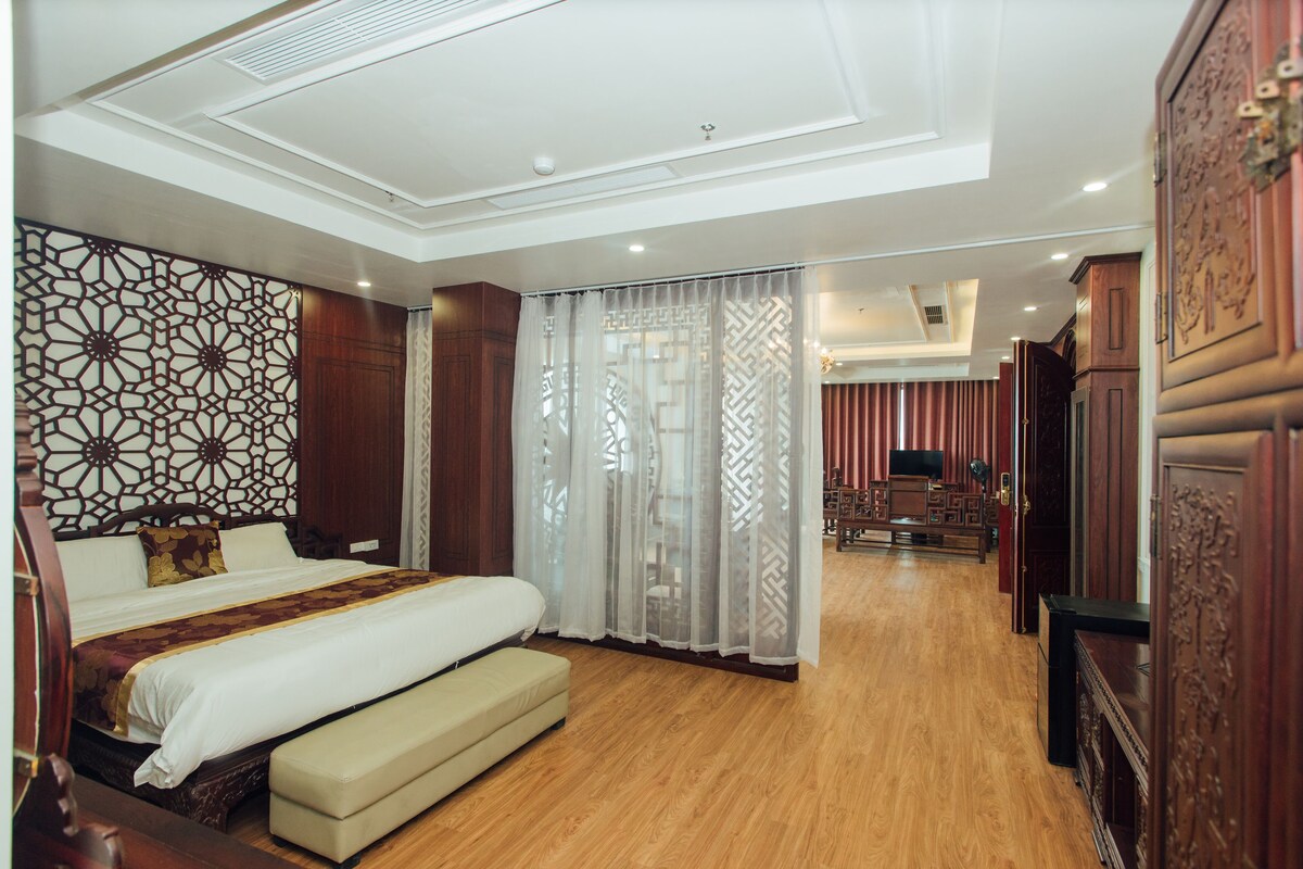 A spacious hotel room with a large bed, a wooden floor, a decorative wall, and a view of the hallway.