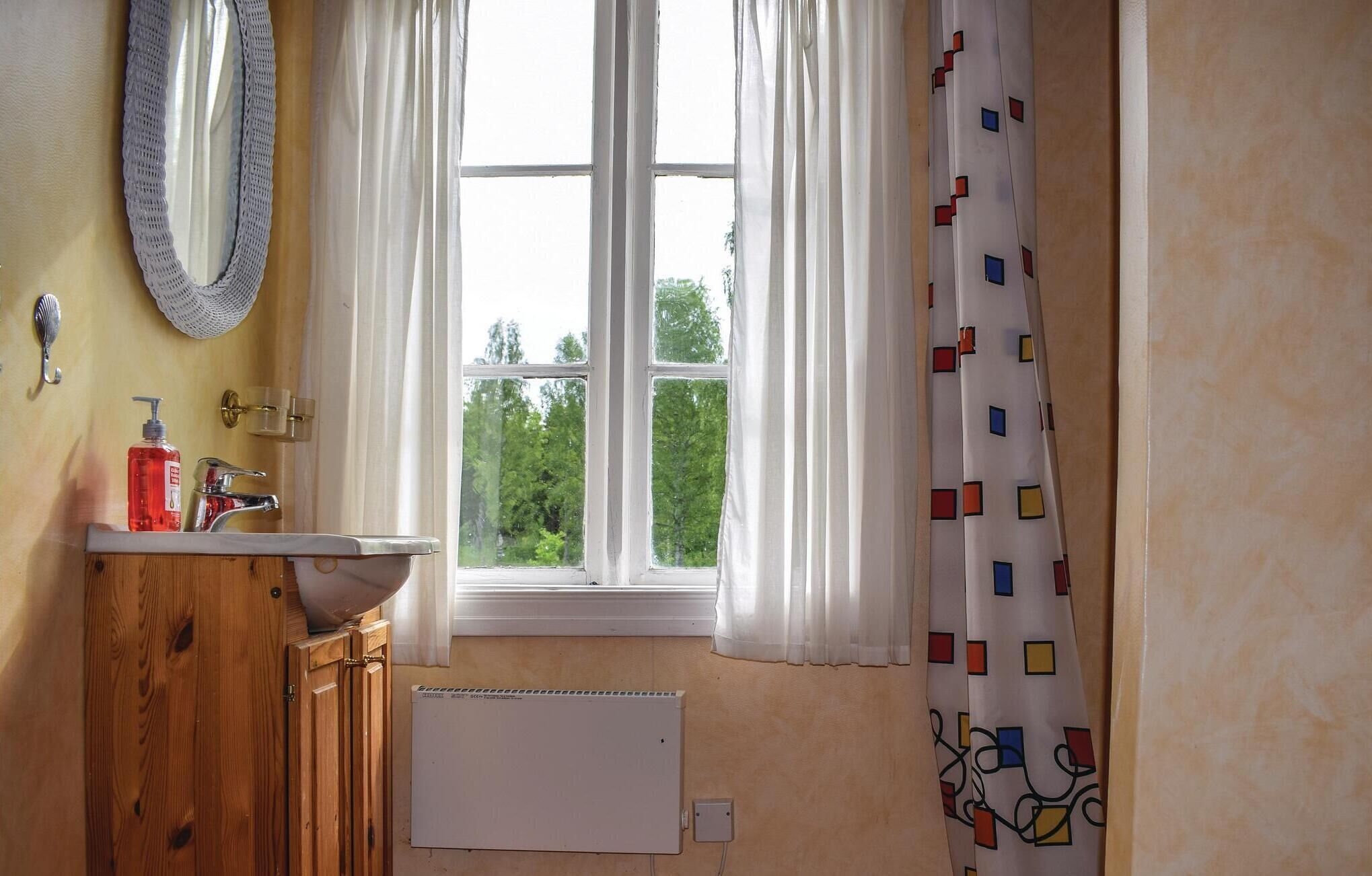 Et badeværelse med en trævådestand, en hvid vask, et spejl, et vindue med hvide gardiner og en radiator.