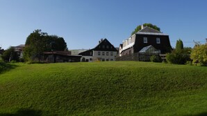 Paesaggio rurale con una grande casa su una collina erbosa, circondata da edifici più piccoli e alberi.
