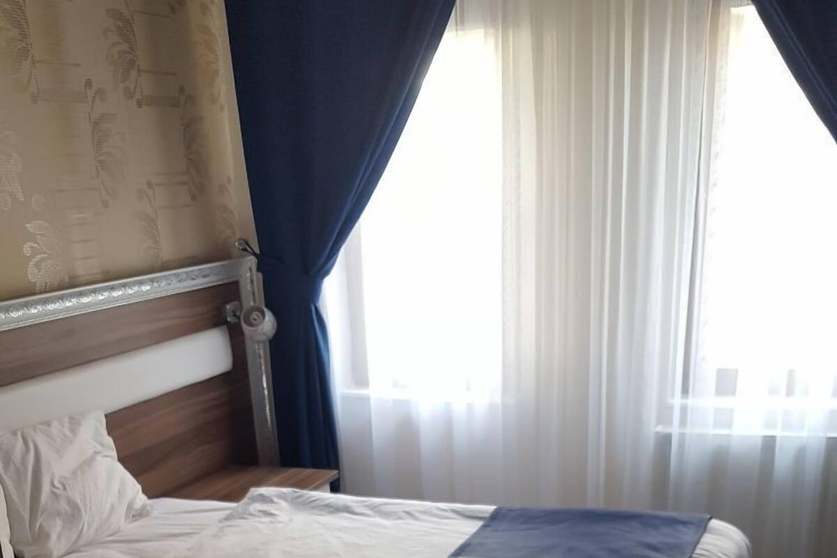 Een net opgemaakt bed met een blauw-witte sprei, een houten hoofdbord en een raam met dunne gordijnen.