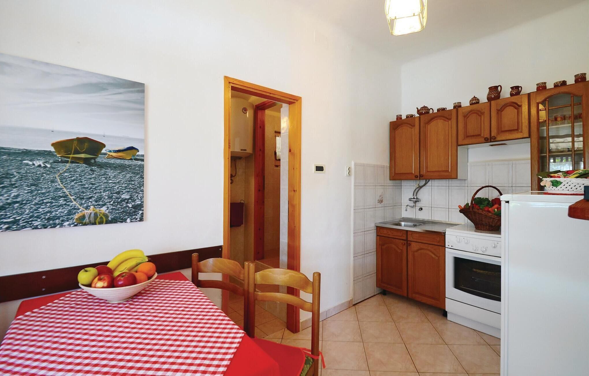 Une cuisine compacte avec des meubles en bois, une cuisinière blanche et un coin repas agrémenté d’une nappe à carreaux et d’un panier de fruits.