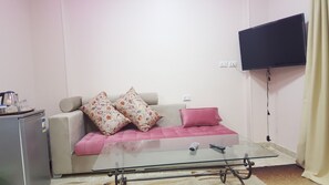 Ein Wohnzimmer mit einer Couch, einem Glastisch, einem an der Wand montierten Fernseher und einer kleinen Küchenzeile mit Wasserkocher und Gläsern.