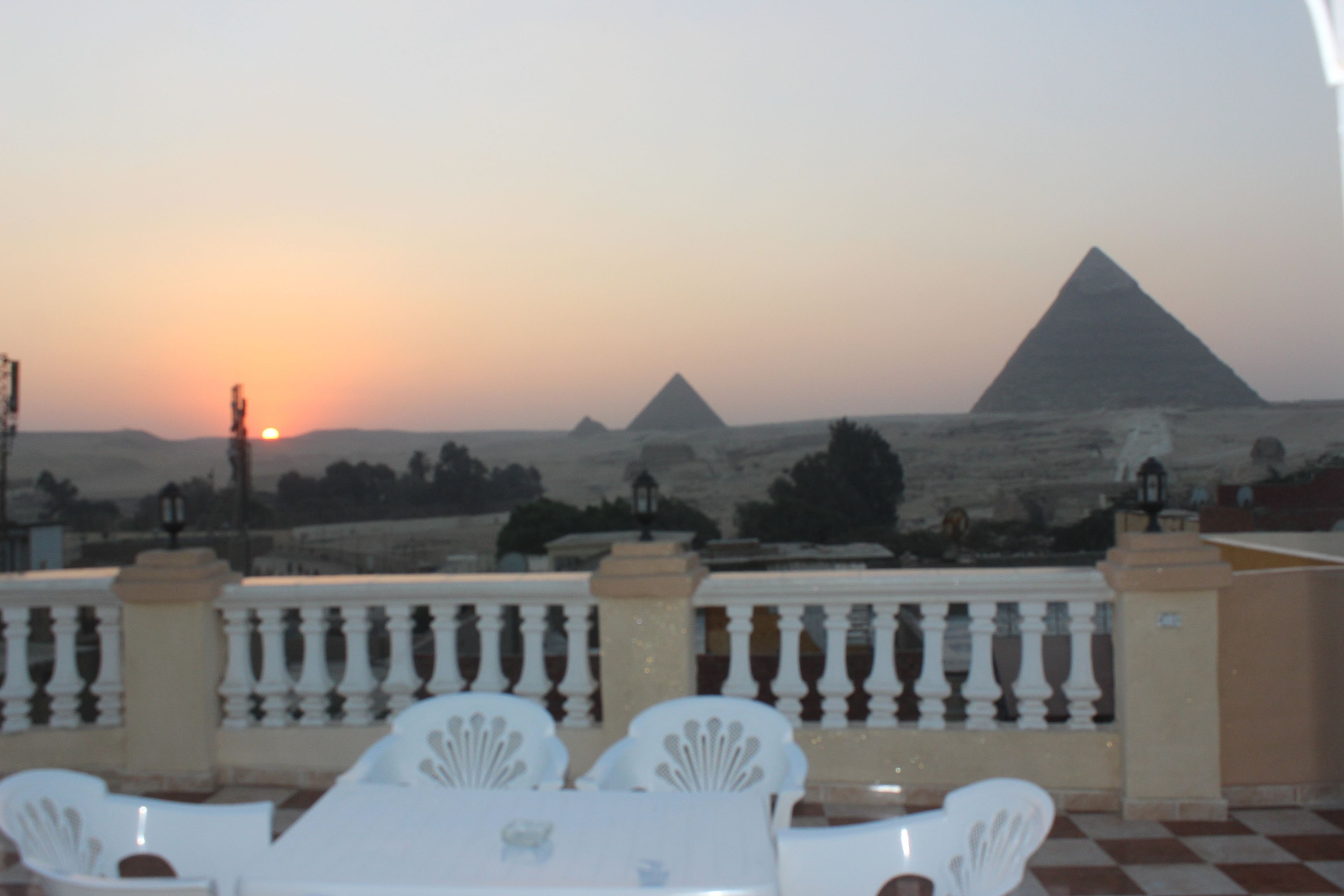 A terrace with a checkered floor, white chairs, and a table. In the background, there are pyramids and a sunset.