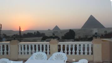 A terrace with a checkered floor, white chairs, and a table. In the background, there are pyramids and a sunset.