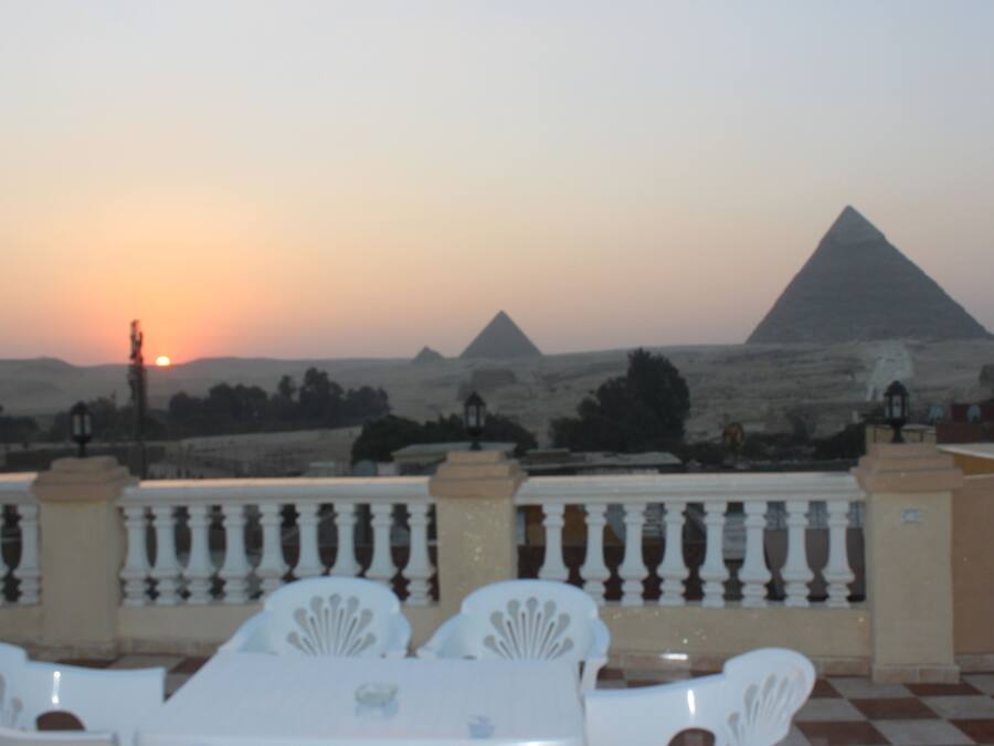 A terrace with a checkered floor, white chairs, and a table. In the background, there are pyramids and a sunset.