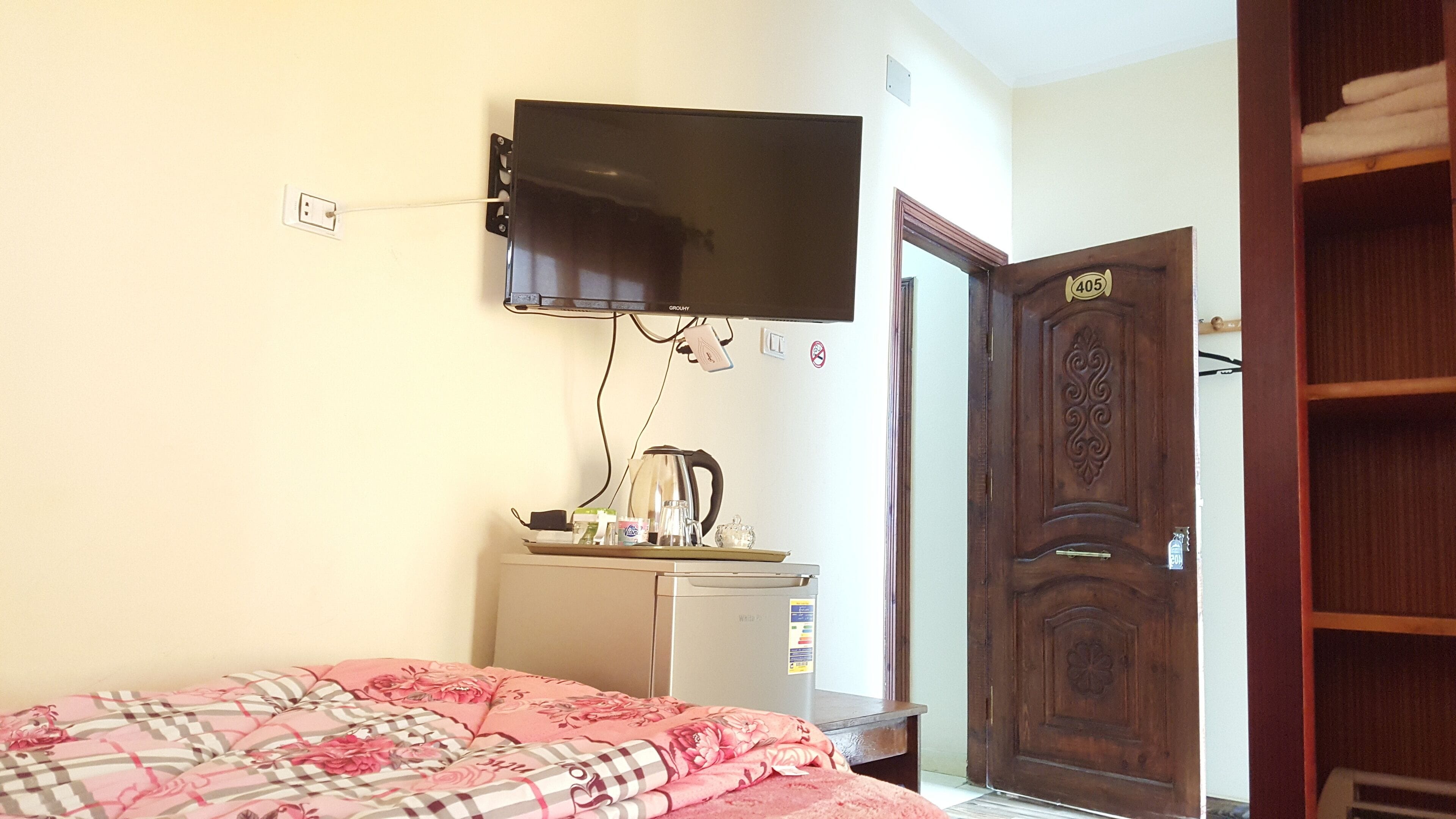 Een kamer met een bed, een aan de muur bevestigde televisie, een kleine koelkast en een houten deur met nummerbord.
