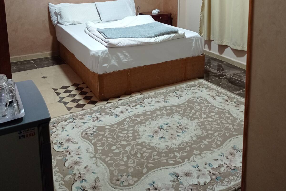 A neatly made bed with white linens, a patterned rug, a nightstand with a lamp, and an air conditioning unit on the wall.
