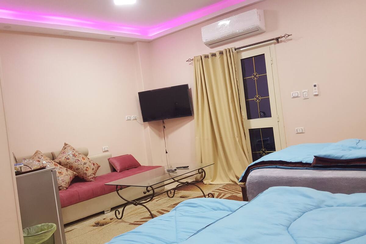A hotel room with a bed, a sofa, a TV mounted on the wall, and an air conditioning unit.