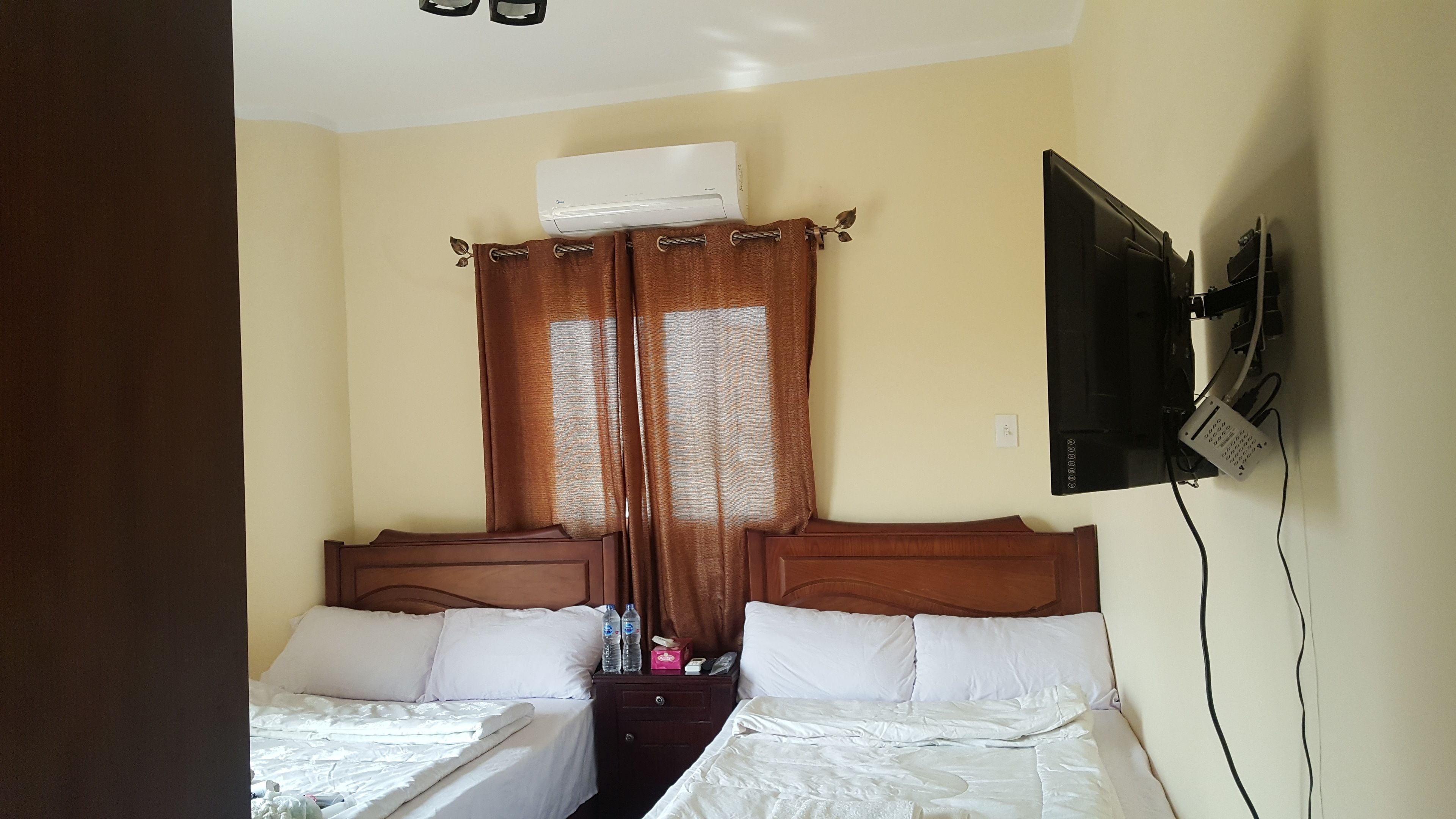 A hotel room with two beds, a TV mounted on the wall, an air conditioning unit above the window, and a bedside table with a bottle and a box.