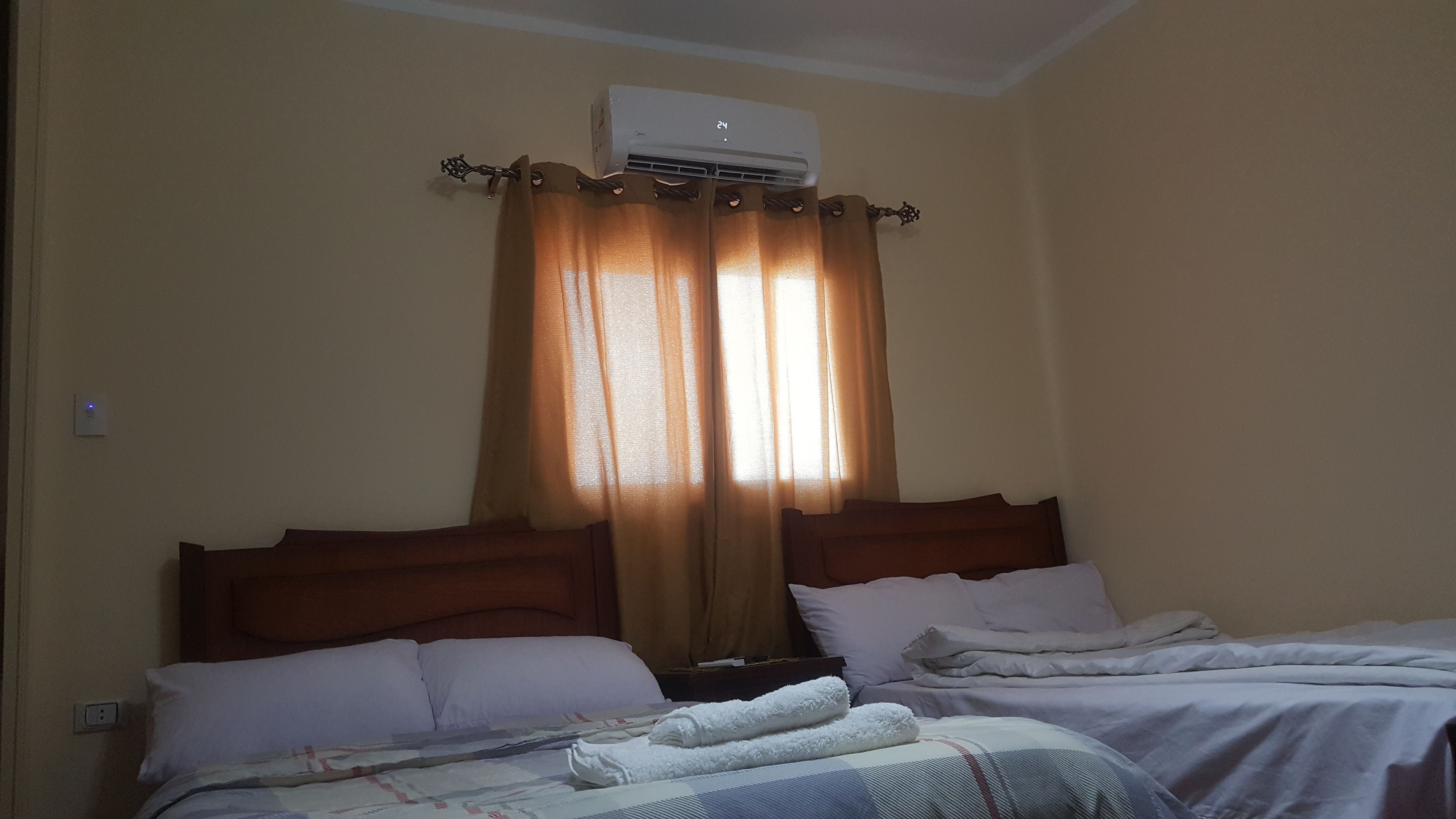 Habitació d'hotel amb dos llits, parets beix i aire condicionat al sostre, i una finestra amb cortines.