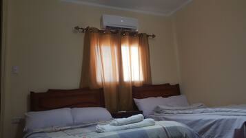 Une chambre d’hôtel avec deux lits, des murs de couleur beige, un climatiseur fixé au plafond et une fenêtre avec des rideaux.