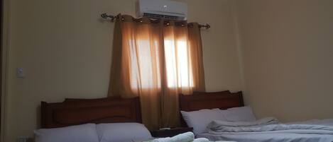 Habitació d'hotel amb dos llits, parets beix i aire condicionat al sostre, i una finestra amb cortines.