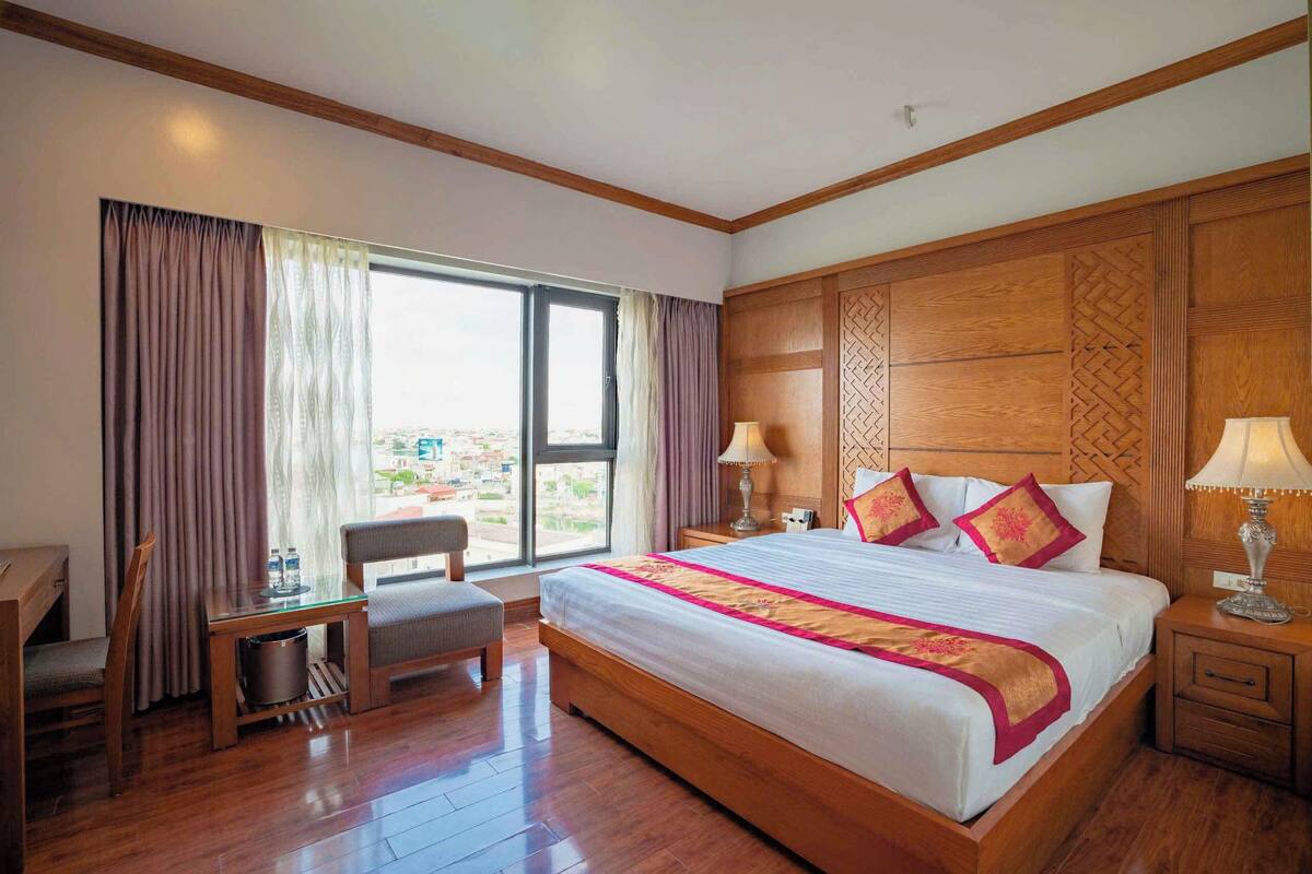 A hotel room with a large bed, wooden headboard, two bedside tables with lamps, a small table with a chair, and a view through the window.