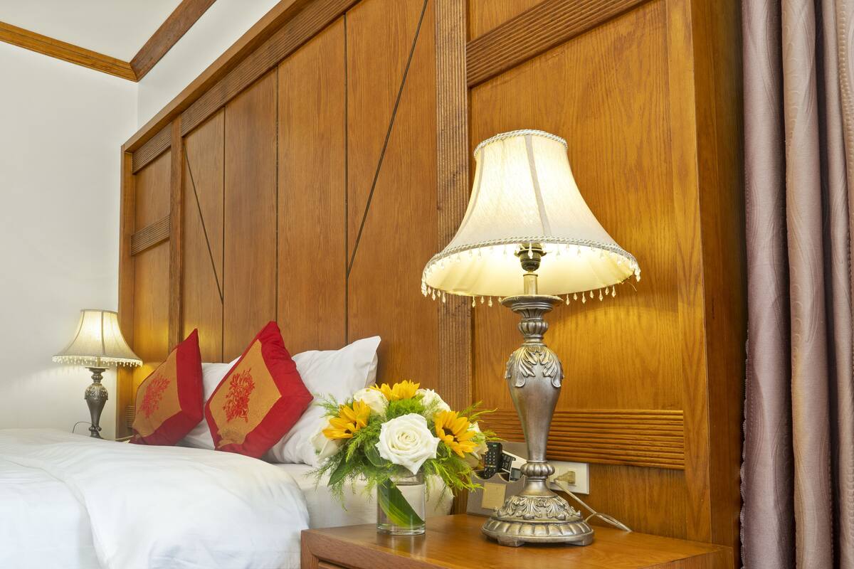 A neatly made bed with white bedding, a bedside lamp, a vase with flowers, and a decorative lamp on a wooden nightstand.