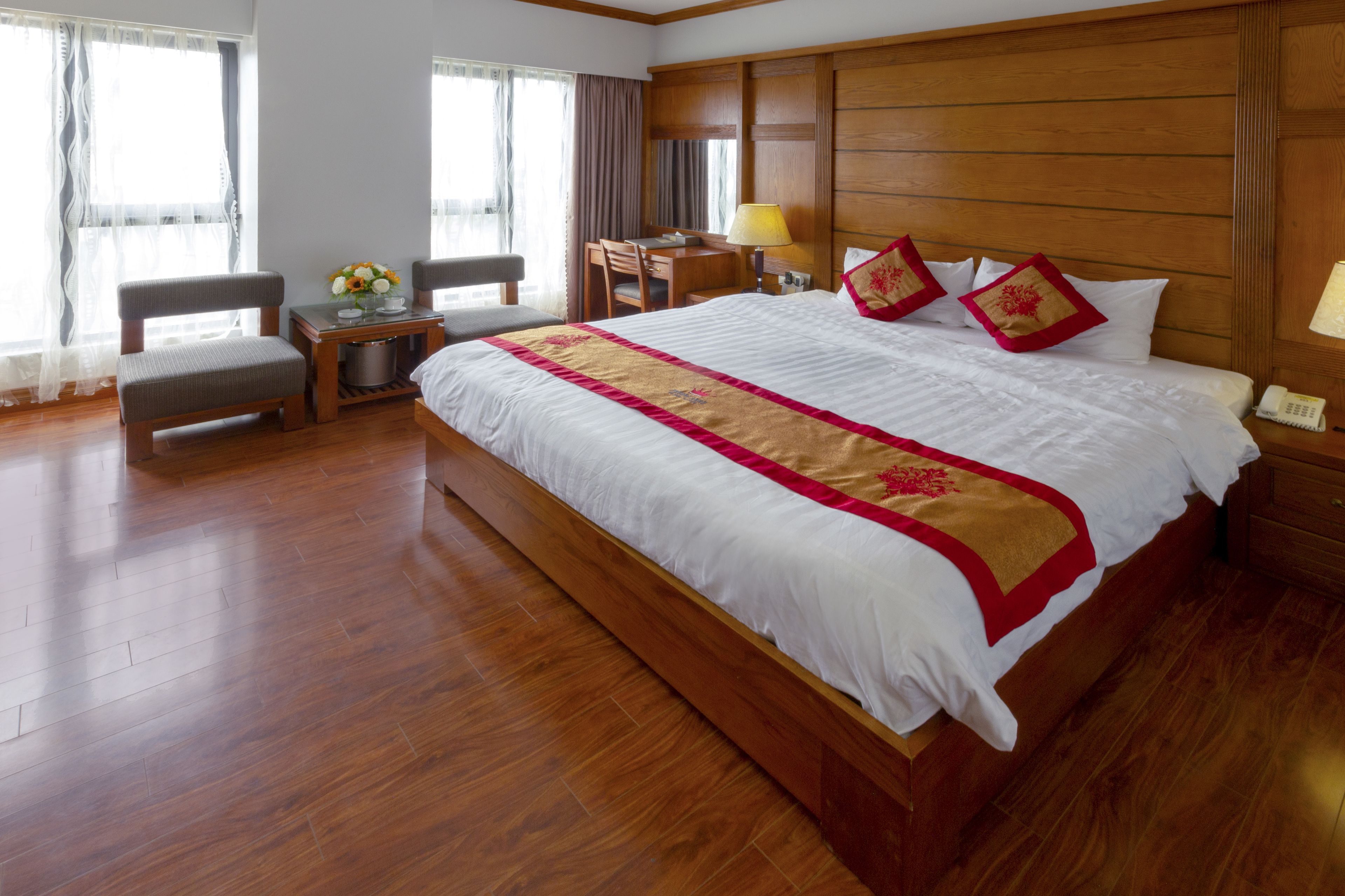 A large bed with a wooden headboard, a bedside table with a lamp, a chair, and a small table with a vase of flowers.