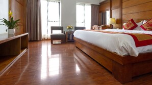A hotel room with a large bed, wooden flooring, a desk with a chair, a small table with a lamp, and a potted plant.