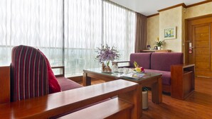 A hotel room with a wooden bench, a sofa, a glass coffee table, and a large window with sheer curtains.