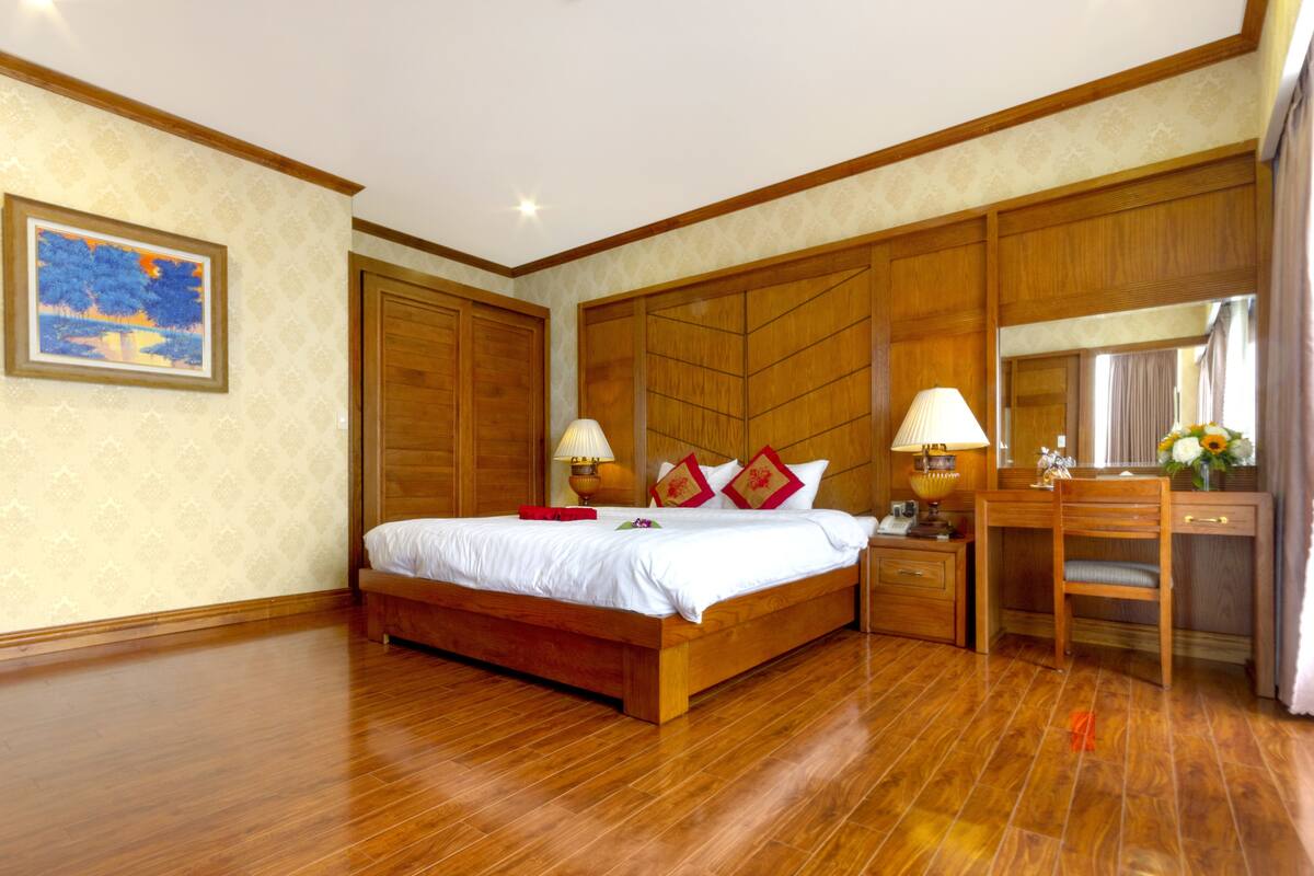 A spacious bedroom with a large bed, wooden headboard, bedside tables with lamps, a desk, and a mirror.